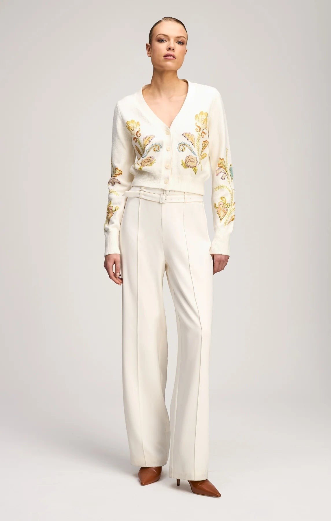 Model wearing a Coats and co. cream V-neck cardigan with intricate floral embroidery, paired with white wide-leg trousers.